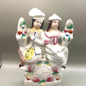 Puede incluir: Un reloj de sobremesa de cerámica blanca con dos figuras que llevan sombreros, sentadas una al lado de la otra. La esfera del reloj está en la base con números negros del 1 al 12 y dos manecillas. Las figuras tienen detalles en pintura amarilla, rosa y verde.