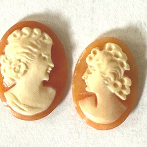 May include: Two oval-shaped cameos featuring cream-colored profiles of individuals with curly hair set against a translucent, warm orange-brown background. Each piece displays a detailed relief carving of a head and shoulders facing to the right.