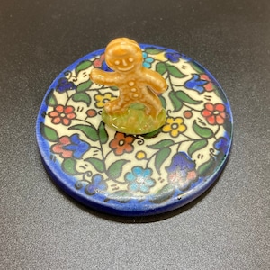 May include: A ceramic gingerbread man figurine standing on a round, blue and white ceramic tile with a floral pattern.
