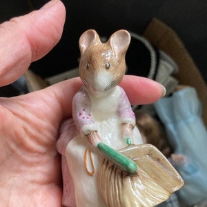 Könnte beinhalten: Eine kleine Keramikfigur einer Maus, die ein lila Kleid trägt und einen grünen Besen hält. Die Maus sitzt auf einem braunen und weißen Korb.
