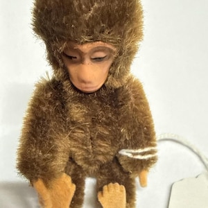 May include: A small, seated monkey figurine covered in brown, textured faux fur. The figure has a smooth, light tan face with closed eyes and matching tan felt-like hands and feet. A white string is attached to the side of the monkey's arm.