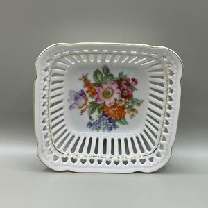 Op de afbeelding: Een vierkante witte porseleinen schaal met een delicate opengewerkte rand en gouden afwerking. Het midden van de schaal toont een kleurrijk bloemstuk met roze, oranje, paarse en blauwe bloemen, omringd door groene bladeren en stengels.