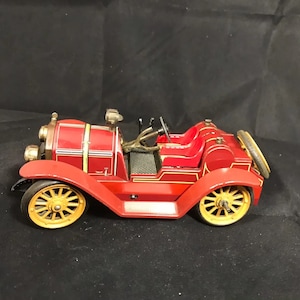 Può includere: Un'auto giocattolo vintage rossa con dettagli dorati e ruote nere. L'auto ha due posti a sedere e una griglia anteriore dettagliata.