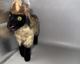 Puede incluir: Un juguete de oveja de peluche vintage con cara, orejas y patas negras. La oveja tiene pelaje marrón claro y ojos verdes. El juguete está en una caja blanca.