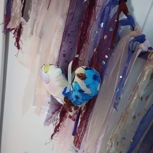 Op de afbeelding: Een handgemaakt hartvormig stoffen ornament met een bloemenpatroon in tinten blauw, wit en bruin. Het ornament hangt aan een wit lint en is omgeven door verschillende gekleurde stofstroken.