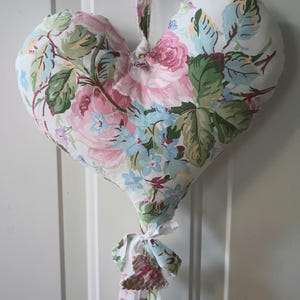 Op de afbeelding: Een hartvormig bloemkussen met een witte achtergrond en roze, blauwe en groene bloemenprint. Het kussen hangt aan een witte deur met een bloemenlint.