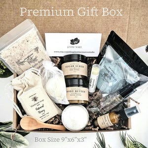 May include: A premium gift box filled with self-care items. The box contains sugar scrub, body butter, bath salts, a wooden spoon, soap, a shower steamer, lip balm, and linen spray. Box dimensions are 9" x 6" x 3".