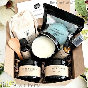 May include: A gift box measuring 6 inches by 6 inches by 4 inches containing eight items: a beige drawstring bag with handmade soap, a shower steamer in a black pouch, a white candle in a black tin, a wooden spoon, body butter, sugar scrub, and linen spray.