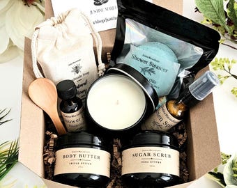 May include: A gift box measuring 6 inches by 6 inches by 4 inches containing eight items: a beige drawstring bag with handmade soap, a shower steamer in a black pouch, a white candle in a black tin, a wooden spoon, body butter, sugar scrub, and linen spray.