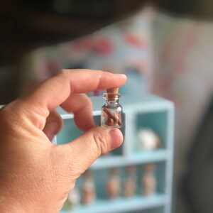 May include: A small, clear glass bottle with a cork stopper, held between fingers. The bottle contains small, brown objects. The background is blurred, with a light blue shelf unit visible.