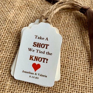May include: White wedding favor tags with brown text that reads "Take a Shot We Tied the Knot!" with a red heart and the names "Jonathan & Valeria" and the date "8.15.20".