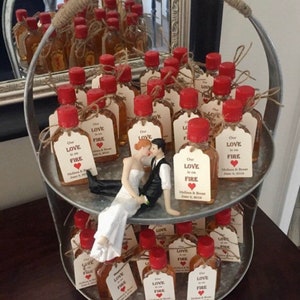 May include: Two-tiered metal stand filled with small glass bottles with red lids and brown liquid. Each bottle has a small tag attached with the text "Our Love is on Fire" and a red heart. A small bride and groom figurine is sitting on the top tier of the stand.
