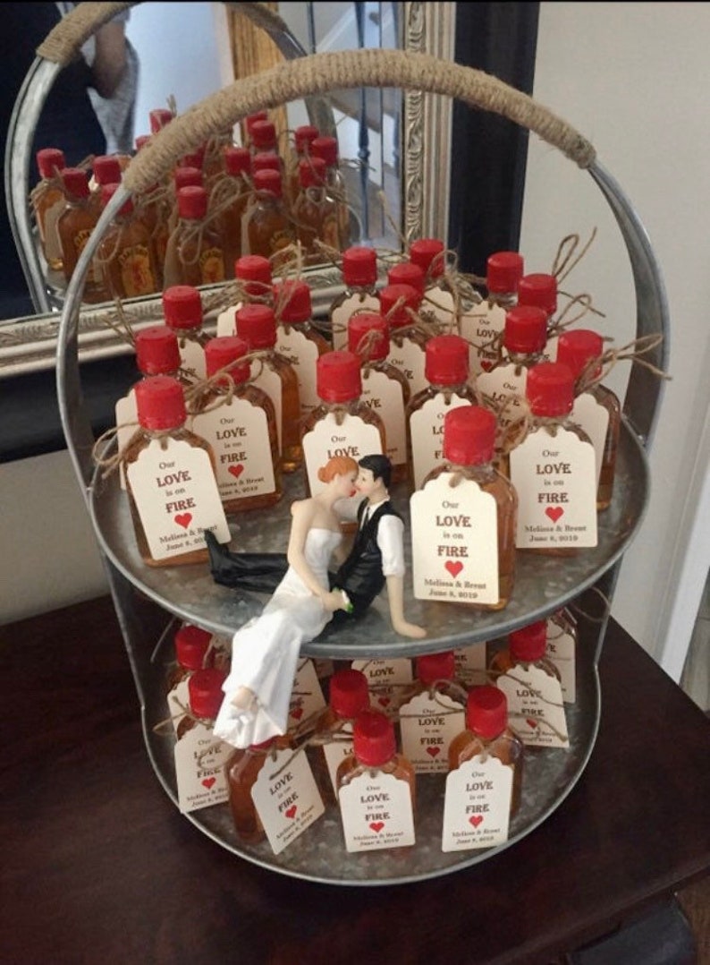 May include: Two-tiered metal stand filled with small glass bottles with red lids and brown liquid. Each bottle has a small tag attached with the text "Our Love is on Fire" and a red heart. A small bride and groom figurine is sitting on the top tier of the stand.