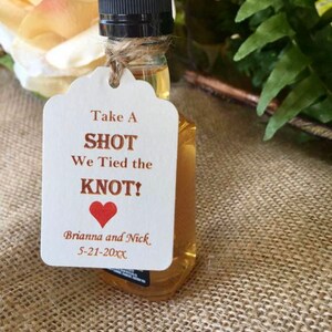 May include: A small bottle of liquor with a white tag attached. The tag says "Take A Shot We Tied the Knot!" with a red heart and the names Brianna and Nick and the date 5-21-20XX.