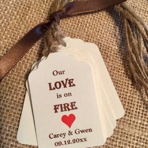 May include: A stack of white wedding favor tags with a brown ribbon and twine. The tags have the text "Our Love is on Fire" with a red heart and the names "Carey & Gwen" and the date "09.12.20XX".