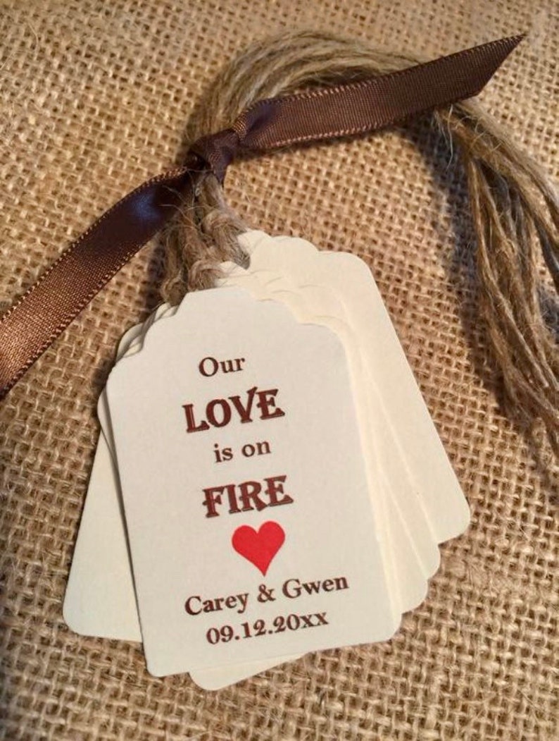 May include: A stack of white wedding favor tags with a brown ribbon and twine. The tags have the text "Our Love is on Fire" with a red heart and the names "Carey & Gwen" and the date "09.12.20XX".
