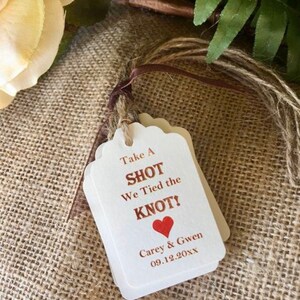 May include: White wedding favor tags with brown twine. The tags read "Take a Shot We Tied the Knot!" with a red heart and the names "Carey & Gwen" and the date "09.12.20XX".