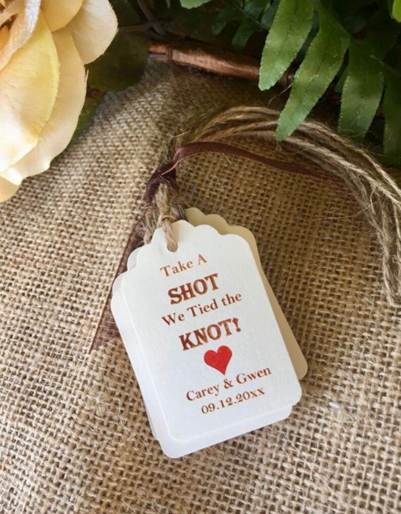May include: White wedding favor tags with brown twine. The tags read "Take a Shot We Tied the Knot!" with a red heart and the names "Carey & Gwen" and the date "09.12.20XX".