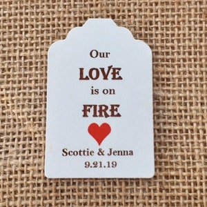 May include: White wedding favor tag with brown text that reads "Our LOVE is on FIRE" with a red heart below the text. The tag also includes the names "Scottie & Jenna" and the date "9.21.19".