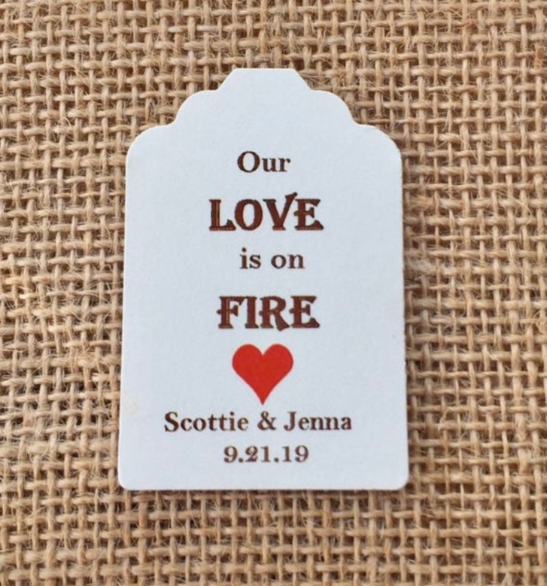May include: White wedding favor tag with brown text that reads "Our LOVE is on FIRE" with a red heart below the text. The tag also includes the names "Scottie & Jenna" and the date "9.21.19".