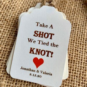 May include: White wedding favor tags with brown text that reads "Take a Shot We Tied the Knot!" with a red heart and the names Jonathan & Valeria and the date 8.15.20.