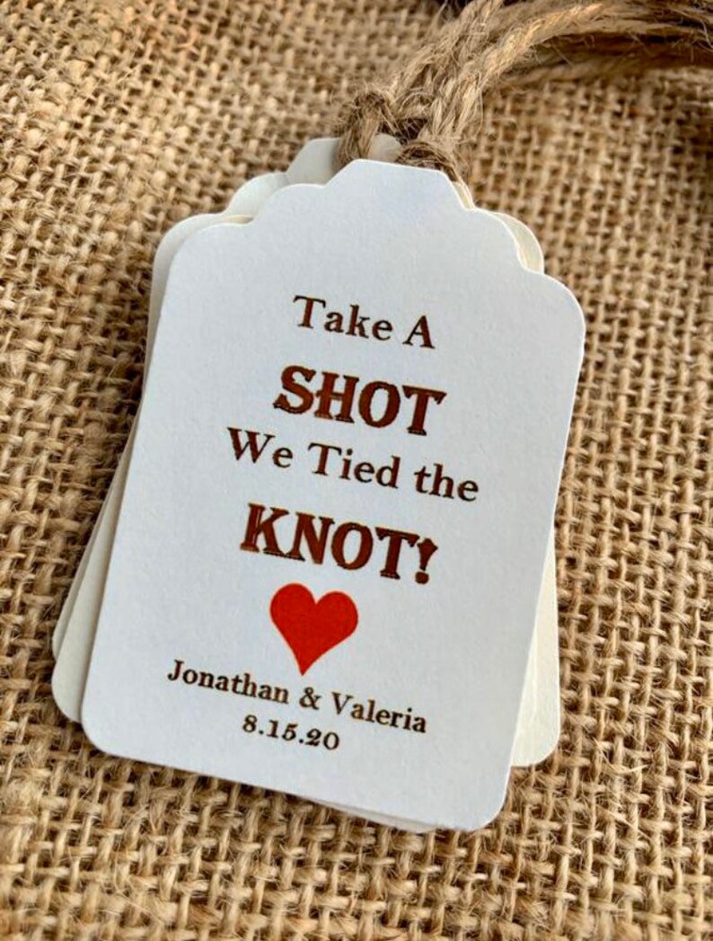 May include: White wedding favor tags with brown text that reads "Take a Shot We Tied the Knot!" with a red heart and the names Jonathan & Valeria and the date 8.15.20.