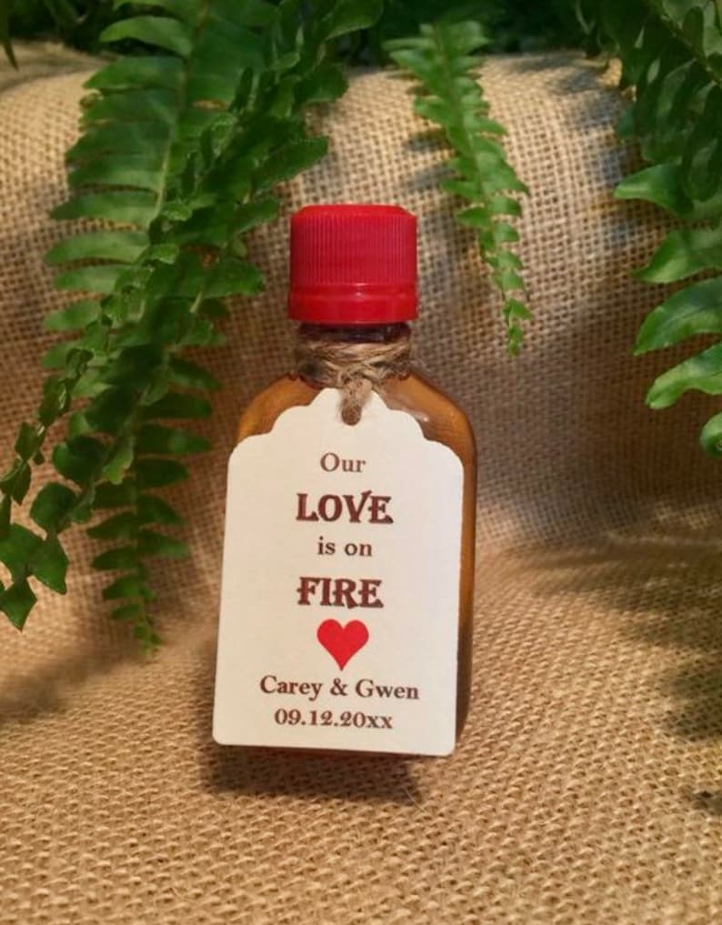May include: A small glass bottle with a red cap and a white label. The label says "Our Love is on Fire" with a red heart and the names "Carey & Gwen" and the date "09.12.20XX".
