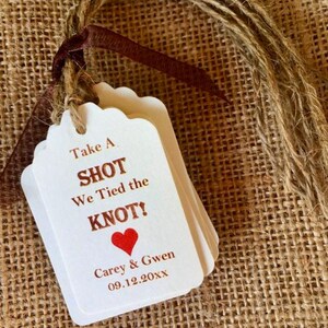 May include: White wedding favor tag with brown text that reads "Take A Shot We Tied the Knot!" with a red heart. The tag also includes the names "Carey & Gwen" and the date "09.12.20XX".