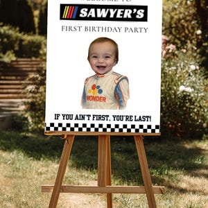 Könnte beinhalten: Eine Holzstaffelei hält ein weißes Schild mit Rennsport-Thema. Auf dem Schild steht "Willkommen zu Sawyers erstem Geburtstag" mit einem lächelnden Kind in einem Rennanzug. Der Text "If you ain't first, you're last!" ist ebenfalls enthalten.