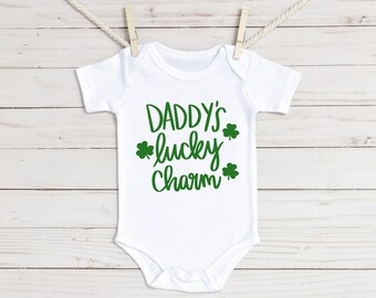 May include: White baby onesie with the text "DADDY'S lucky Charm" in green script and shamrock graphics. The onesie is hanging from a clothesline with wooden clothespins against a white wood background.