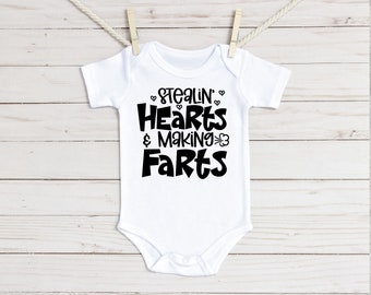 May include: White baby onesie with black text that reads "Stealin' Hearts & Making Farts".