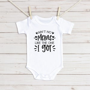 May include: White baby bodysuit with black text: "Ain't No Meme Like The One I Got." The garment is suspended from a clothesline with wooden pegs. The backdrop is light-coloured wooden planks.