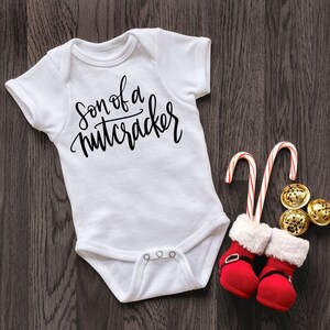 Op de afbeelding: Witte babybody met de tekst "Son of a Nutcracker" in zwart script. De body wordt gecombineerd met rood-witte zuurstokken, miniatuur rode laarsjes met witte rand en gouden belletjes. Een feestelijk vakantiethema.