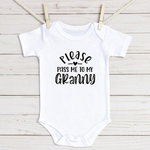 Op de afbeelding: Witte baby romper met zwarte tekst die "Geef me alstublieft aan mijn oma" zegt met een klein hartje boven het woord "mijn".