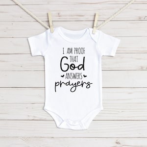 Könnte beinhalten: Weißer Baby-Strampler mit schwarzem Text, der "Ich bin der Beweis, dass Gott Gebete erhört" lautet.