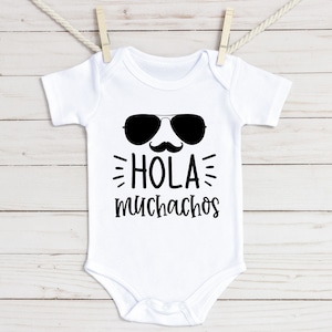 May include: White baby onesie with black text that says "Hola muchachos" with a pair of sunglasses and a mustache above the text.