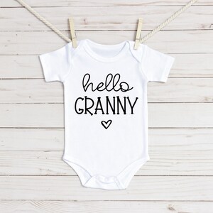 Może przedstawiać: Biały śpioszek dla niemowlęcia z czarnym napisem "hello granny" z małym sercem pod tekstem.