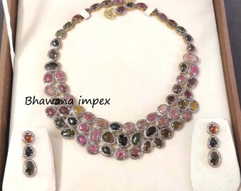 May include: A multi-colored gemstone necklace and earrings set. The necklace features a layered design with oval-shaped gemstones in shades of pink, green, and black. The earrings are a matching set with three oval-shaped gemstones.