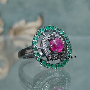 May include: A silver ring with a pink gemstone surrounded by a circle of green gemstones and smaller white gemstones.