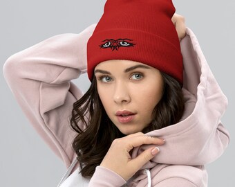 May include: Red knit beanie with a black embroidered design of two eyes with tribal-style accents.