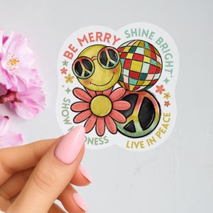 May include: A white sticker with a smiley face wearing peace sign sunglasses, a disco ball, a peace sign, and a flower. The sticker has the phrases "Be Merry", "Shine Bright", "Show Kindness", and "Live in Peace".