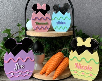 May include: Four Easter egg-shaped decorations with Mickey Mouse ears and bows. The eggs are painted in different colors and have names written on them: Joe, Hannah, Aiden, and Nicole.