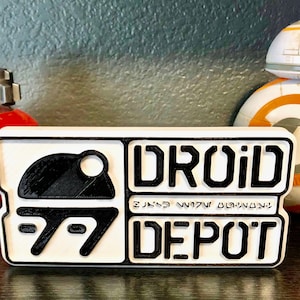 Könnte beinhalten: Ein weißes Schild mit schwarzer Beschriftung, auf dem "DROID DEPOT" steht, mit einem schwarz-weißen Droiden-Symbol. Das Schild steht auf einer Holzoberfläche vor einem weißen und orangefarbenen BB-8-Spielzeug.