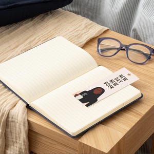 Op de afbeelding: Een open notitieboek met een blauwe kaft en gelinieerde pagina's, met een bladwijzer met een illustratie van een persoon die een mok vasthoudt. De bladwijzer luidt "NEW DAY NEW BOOK". Een blauwe bril ligt op een houten oppervlak.