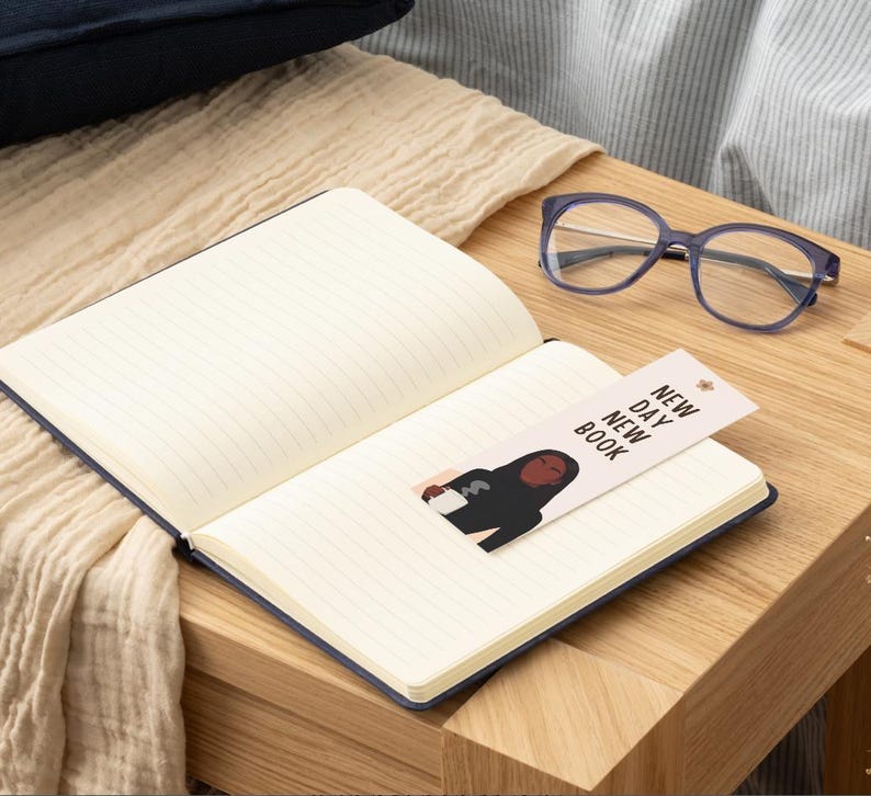 Op de afbeelding: Een open notitieboek met een blauwe kaft en gelinieerde pagina's, met een bladwijzer met een illustratie van een persoon die een mok vasthoudt. De bladwijzer luidt "NEW DAY NEW BOOK". Een blauwe bril ligt op een houten oppervlak.