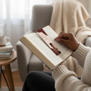 Op de afbeelding: Een bladwijzer met de tekst "Bound and Determined" en een illustratie van een persoon met een hoofddoek is in een open boek geplaatst. De bladwijzer heeft een bordeauxrode en beige kleurstelling. De persoon draagt een cr&egrave;mekleurige trui.