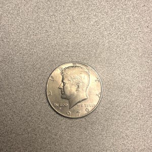 May include: A silver 1970 Kennedy half dollar coin. The coin features a profile of John F. Kennedy and the inscriptions "IN GOD WE TRUST" and "WE TRUST". The year "1970" is also visible.