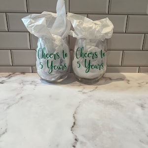 K&ouml;nnte beinhalten: Zwei klare, stielose Weingl&auml;ser mit dem gr&uuml;nen Text "Cheers to 5 Years". Die Gl&auml;ser sind mit wei&szlig;em Seidenpapier gef&uuml;llt. Der Hintergrund ist eine wei&szlig;e Marmorarbeitsplatte und eine wei&szlig;e Fliesenspiegel.