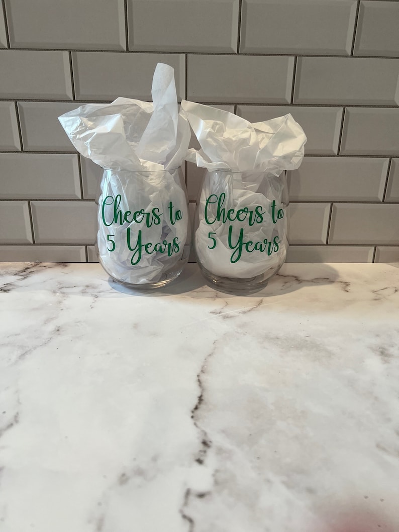 K&ouml;nnte beinhalten: Zwei klare, stielose Weingl&auml;ser mit dem gr&uuml;nen Text "Cheers to 5 Years". Die Gl&auml;ser sind mit wei&szlig;em Seidenpapier gef&uuml;llt. Der Hintergrund ist eine wei&szlig;e Marmorarbeitsplatte und eine wei&szlig;e Fliesenspiegel.