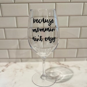 K&ouml;nnte beinhalten: Ein klares Weinglas mit dem Schriftzug "because mommin', ain't easy" in schwarzer Schrift. Das Glas hat einen langen Stiel und eine breite Schale. Hintergrund: wei&szlig;e geflieste Wand und wei&szlig;e Marmorarbeitsplatte.